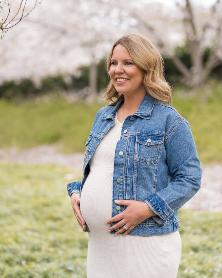 Pregnant mom smiling while holding baby bump in a grass field surrounded by cherry blossoms