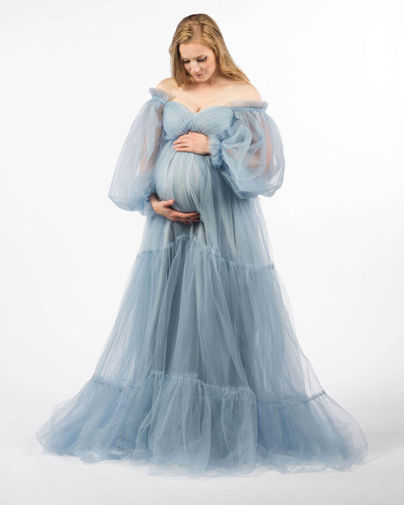 Pregnant mom standing in a white studio wearing a blue flowing gown holding her baby bump with both hands.