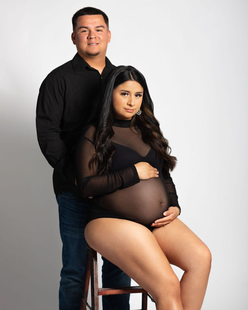 Pregnant wife sitting on stool holding baby bump while husband stands behind her.