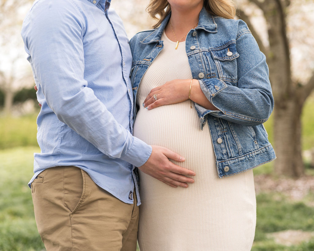 Pregnant woman hugging her husband outdoors while each have one hand on her baby bump.