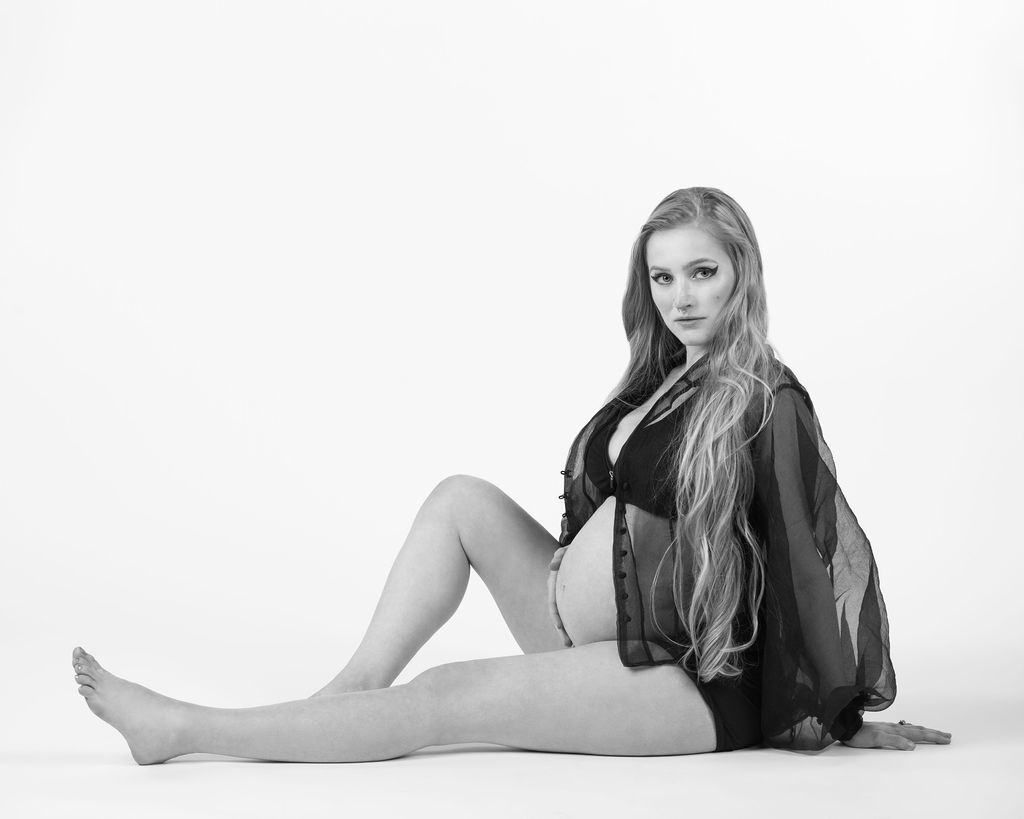 Photo of pregnant woman sitting in a white studio wearing a sheer top while holding her baby bump and looking towards the camera.