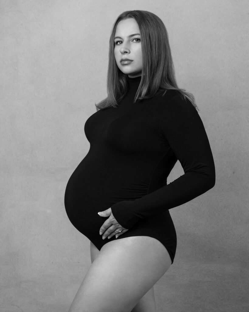 Pregnant mom wearing a black bodysuit looking towards the camera while holding her baby bump with one hand.