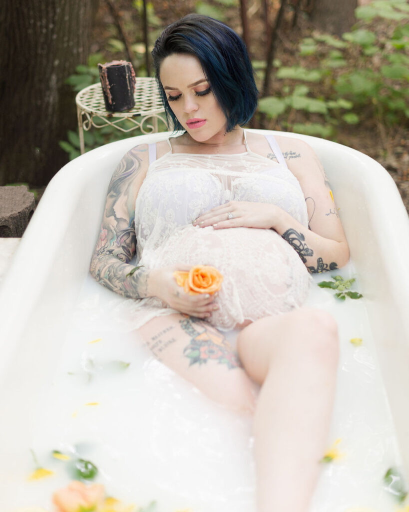 Milk bath photo of a pregnant mom laying in an outdoor bathtub holding a flower yellow flower in one hand and caressing her baby bump with the other hand.