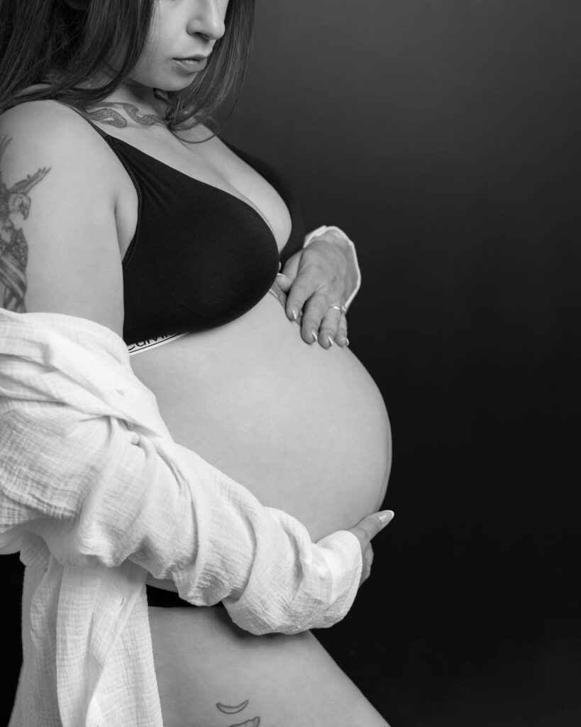 Pregnant woman in black studio wearing a black bra while caressing her baby bump.