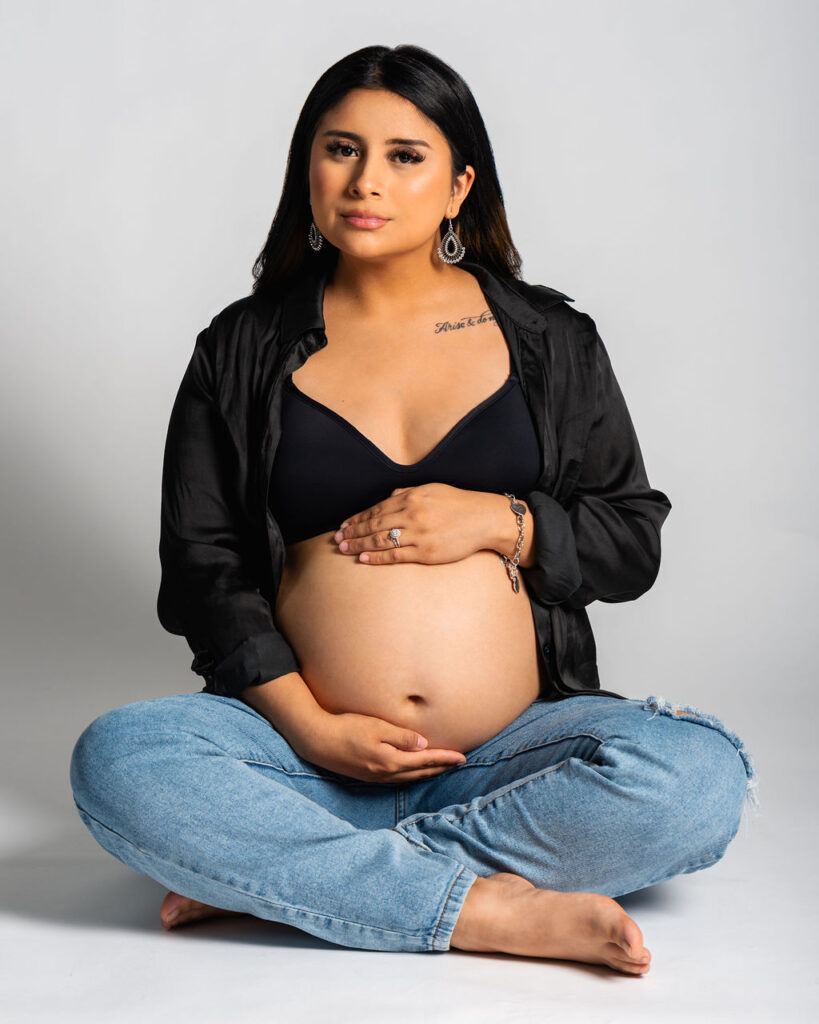 Pregnant woman sitting in a photo studio wearing an unbuttoned black shirt and blue jeans while caressing her baby hump.