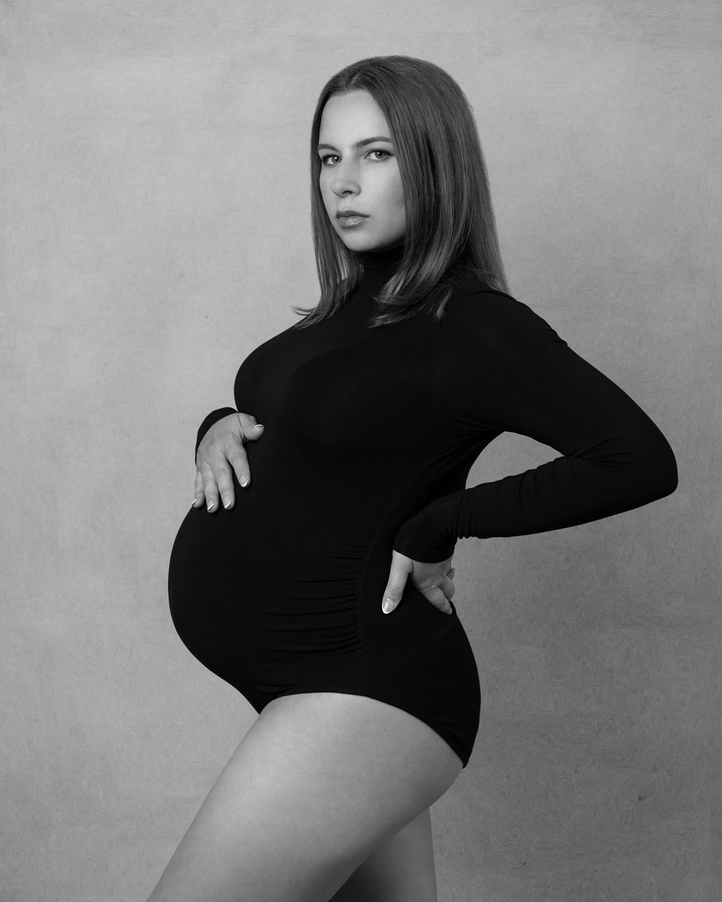 Pregnant woman wearing a black bodysuit posing in a studio holding her baby bump with one hand.