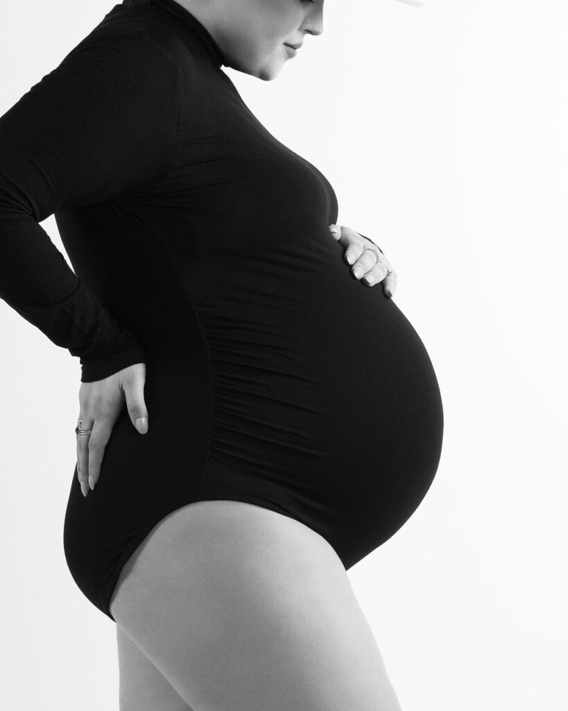 Pregnant woman in a white studio wearing a black long sleeve bodysuit holding her baby bump.