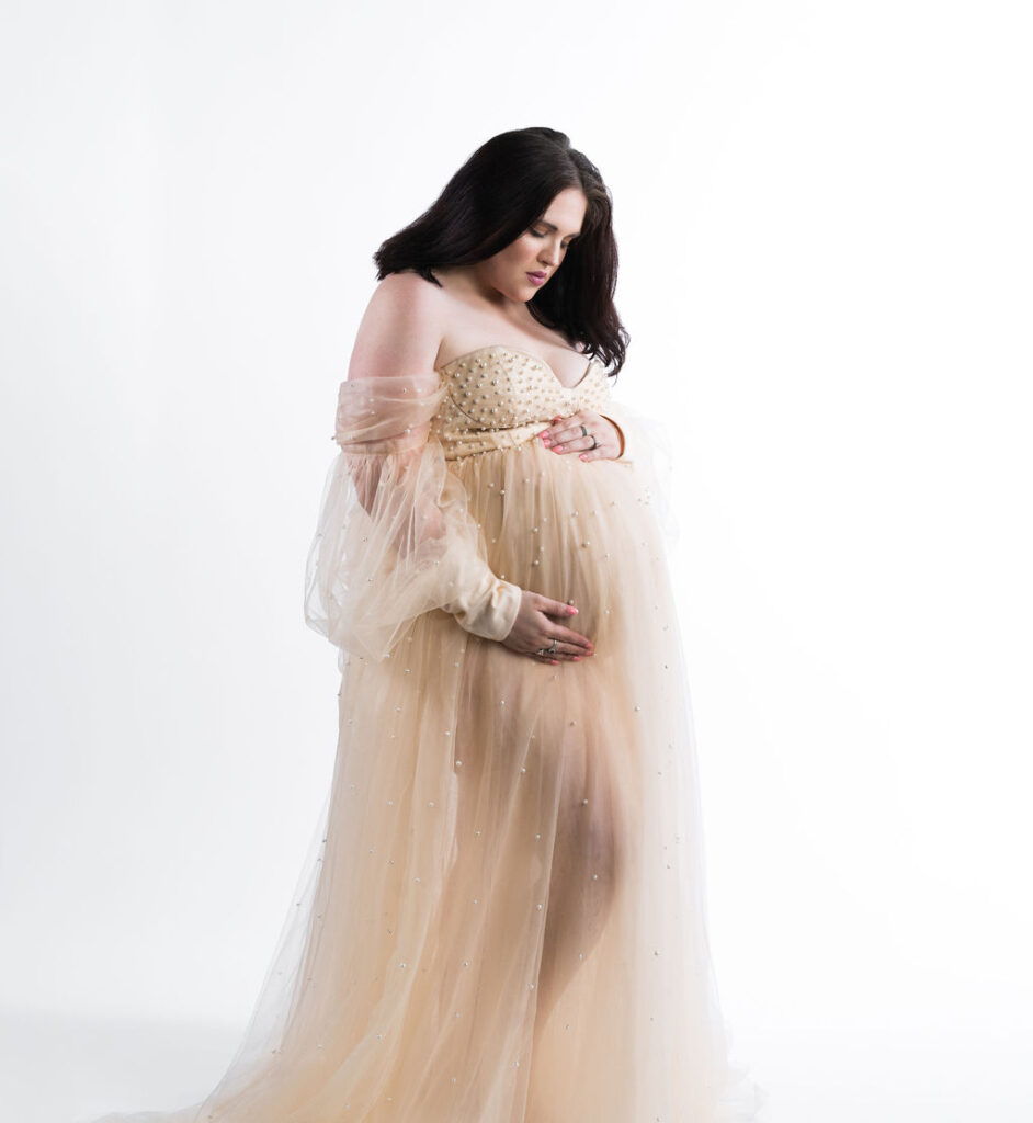 Photo of pregnant woman standing in a white studio with a peach colored sheer gown with pearls on it. The woman is holding her baby bump white looking down at it.