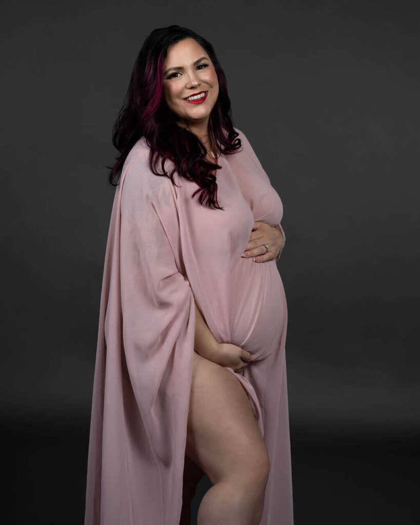 Portrait of pregnant woman standing in a black background wearing a pink flowing fabric while holding her baby bump.