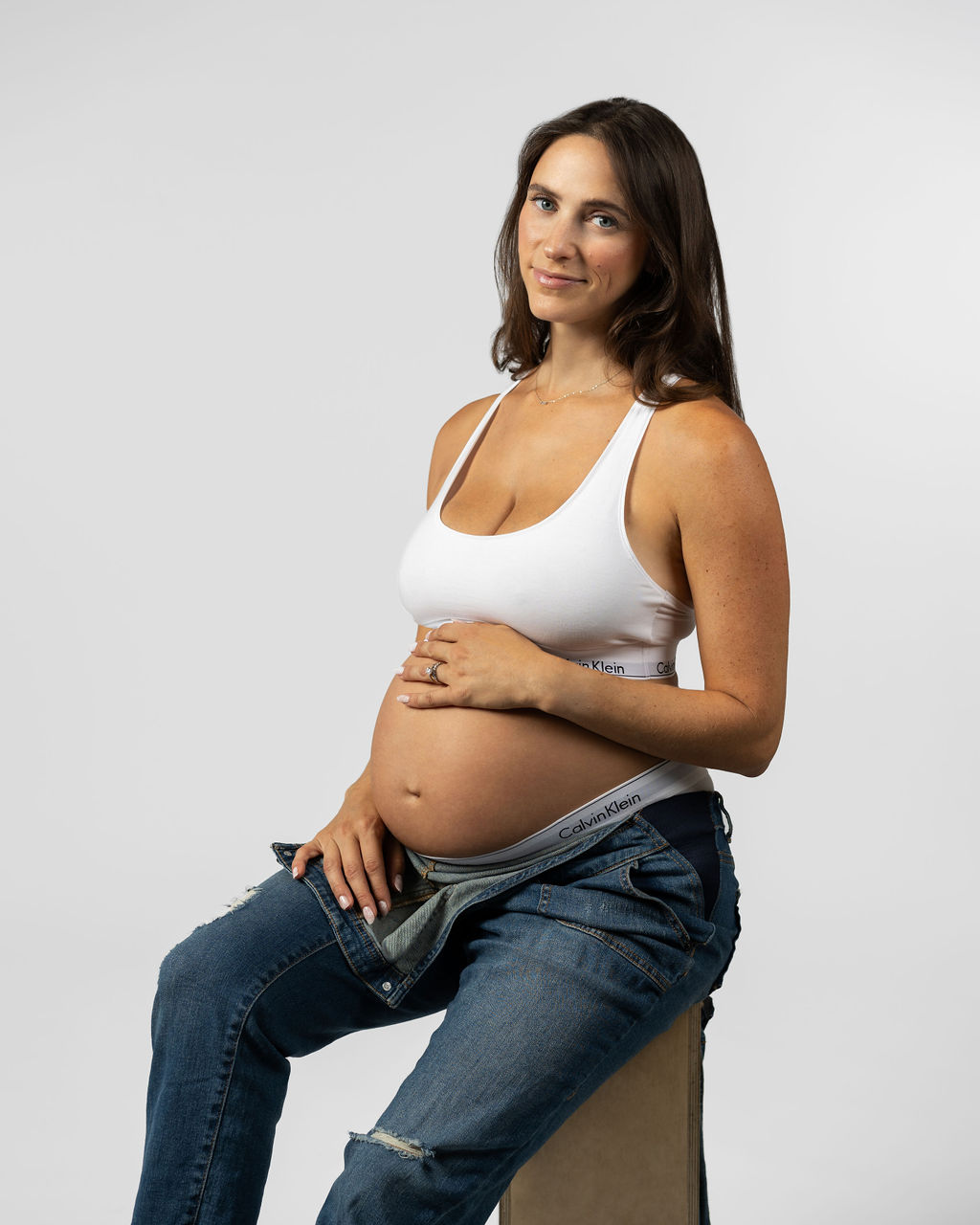 Portrait of a pregnant woman sitting on a wooden box wearing a white bra and blue jeans while smiling at the camera and holding baby bump.