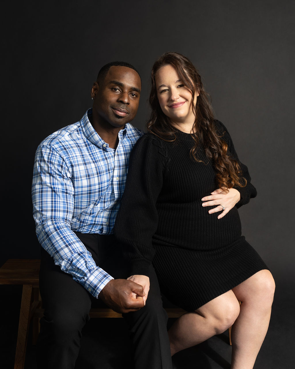 An Austin couple taking maternity photos together in a studio with black background sitting together and holding hands.