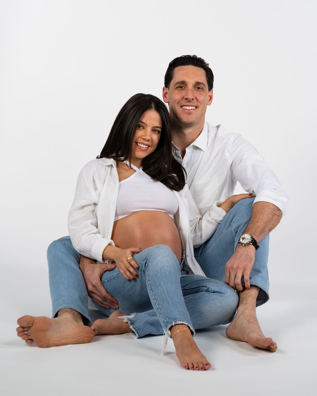 An Austin couple taking maternity photos together in a studio while both wearing blue jeans and a white long sleeve shirt.