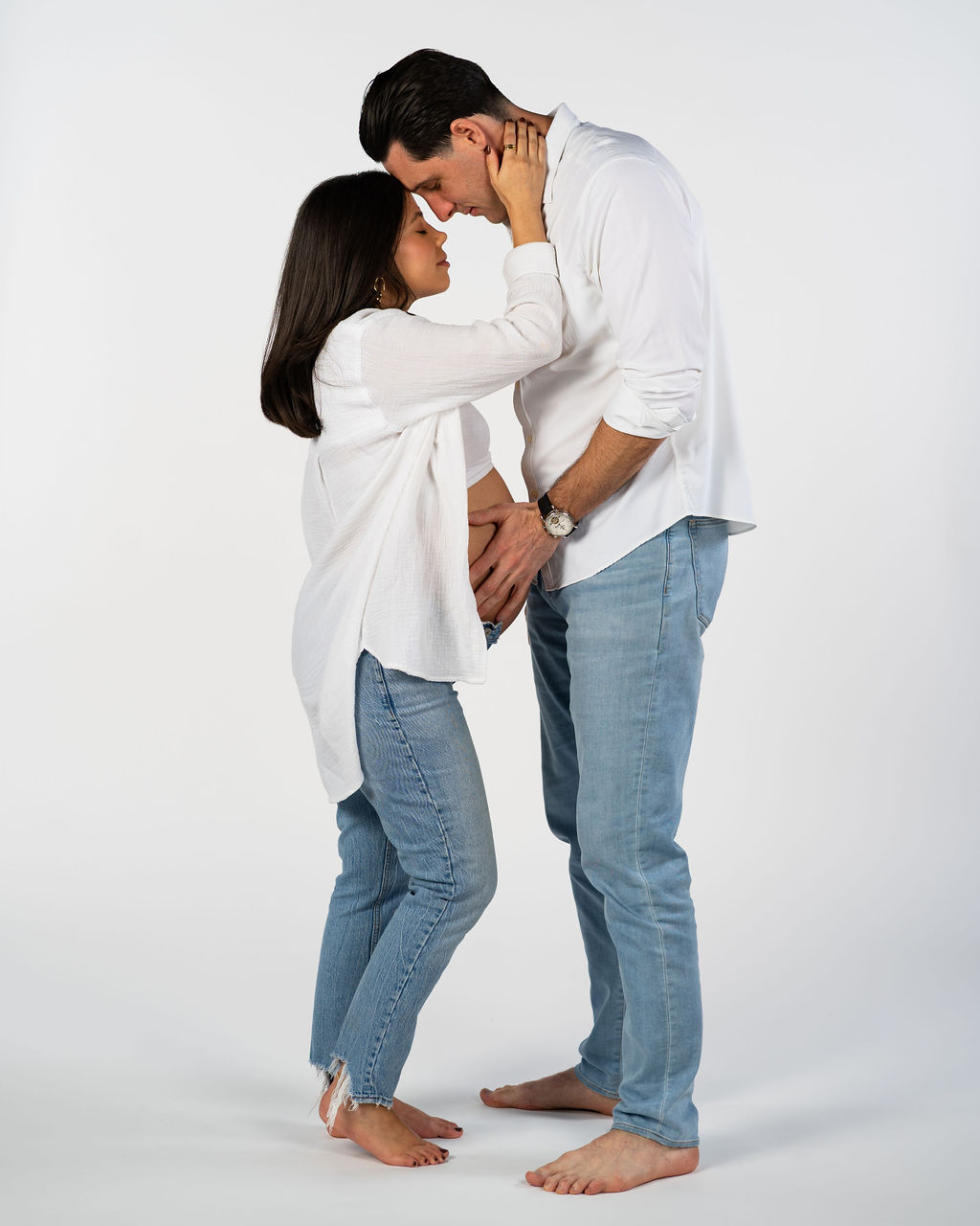 An Austin couple taking maternity photos together in a studio while both wearing blue jeans and a white long sleeve shirt.