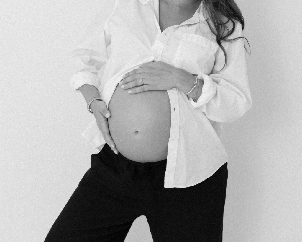 Black-and-white photo of a pregnant woman standing against a plain background, wearing an open button-down shirt and loose pants. She gently holds her belly with both hands.